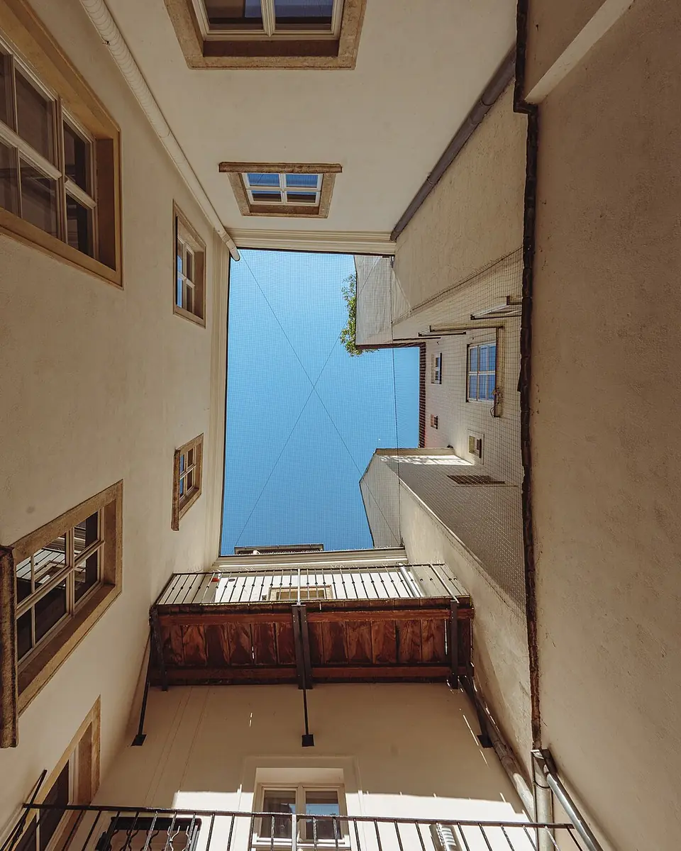 Blick nach oben in einen schmalen Wiener Lichthof mit hellen Putzfassaden, Holzfenstern und einem Stück Himmel